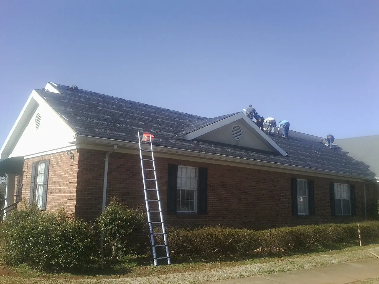 Crew tearing shingles off a brick ranch roof with a ladder set — Emergency Roof Repair work by our crew in Clarks Hill
