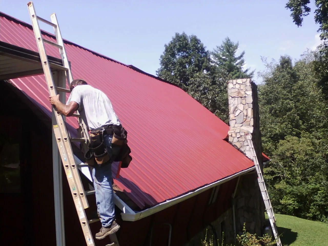 Roofer on a ladder beside a red metal roof and stone chimney — Soffit & Fascia Repair by Roofing Brothers in Clarks Hill